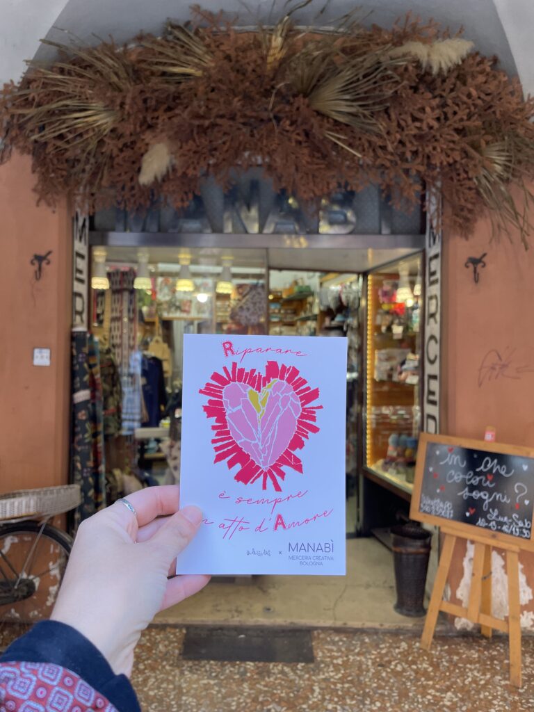 mano che tiene il flyer della merceria con il disegno di un cuore davanti alla bottega sotto i portici di Bologna